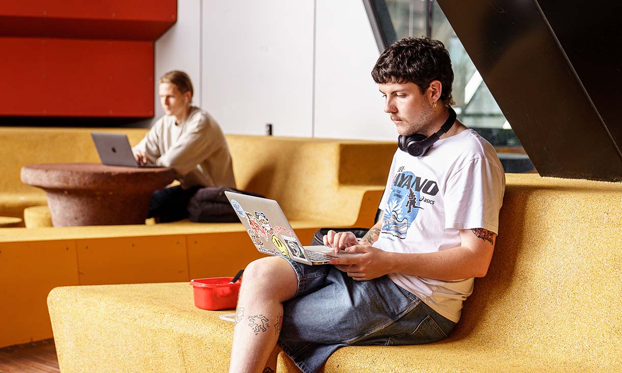 A student sitting in a common area with a laptop, headphones around their neck